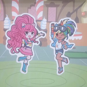 May include: Two cartoon characters, one pink with a cupcake on her shirt and the other rainbow-colored with goggles, are shown in a playful pose. They are both wearing colorful outfits and have big eyes. The characters are on a green background with a brown and white striped background behind them.