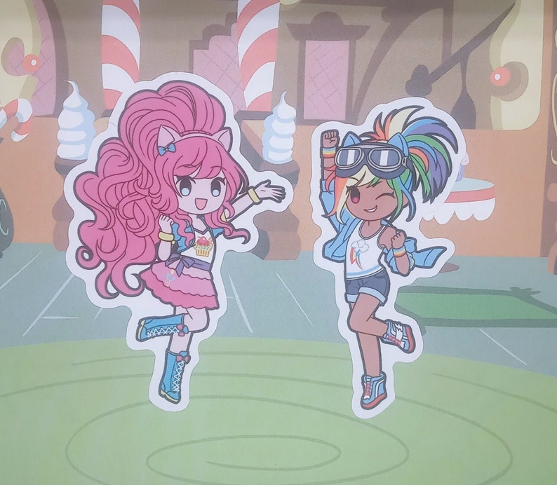 May include: Two cartoon characters, one pink with a cupcake on her shirt and the other rainbow-colored with goggles, are shown in a playful pose. They are both wearing colorful outfits and have big eyes. The characters are on a green background with a brown and white striped background behind them.