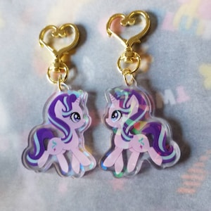 May include: Two iridescent purple and pink pony keychains with gold heart-shaped clasps. The ponies have blue eyes and purple and blue manes and tails. The keychains are a sparkly, holographic material.