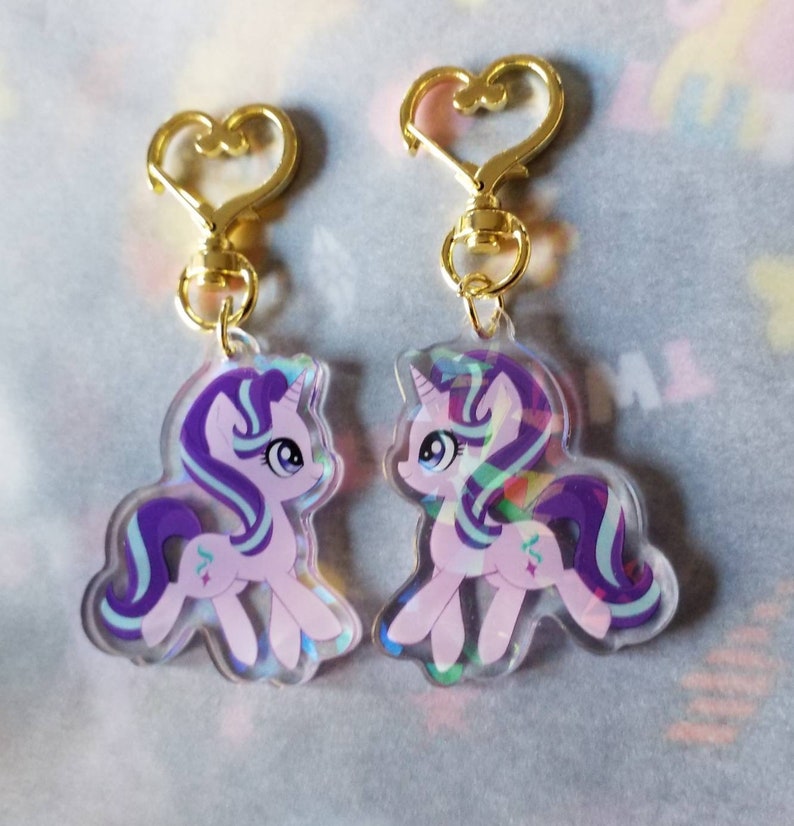 May include: Two iridescent purple and pink pony keychains with gold heart-shaped clasps. The ponies have blue eyes and purple and blue manes and tails. The keychains are a sparkly, holographic material.