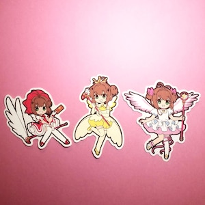 May include: Three anime-style stickers of a girl in different outfits. The stickers feature a girl with brown hair and green eyes, wearing a red, yellow, and white dress, each holding a staff. The background is pink.