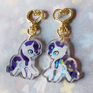 May include: Two iridescent clear acrylic keychains featuring a cartoon image of Rarity from My Little Pony. The keychains have gold heart-shaped clasps.