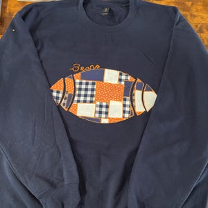 May include: A navy blue crewneck sweatshirt featuring a large, central football-shaped applique made of various fabric patches in orange, white, and navy blue plaid and polka dot patterns. The word Bears is embroidered in orange thread above it.