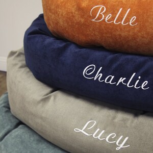 May include: A stack of four round pet beds in different colours and fabrics. The top bed is orange, the second is navy blue, the third is beige, and the bottom is teal. Each bed has a white embroidered name: Belle, Charlie, Lucy.