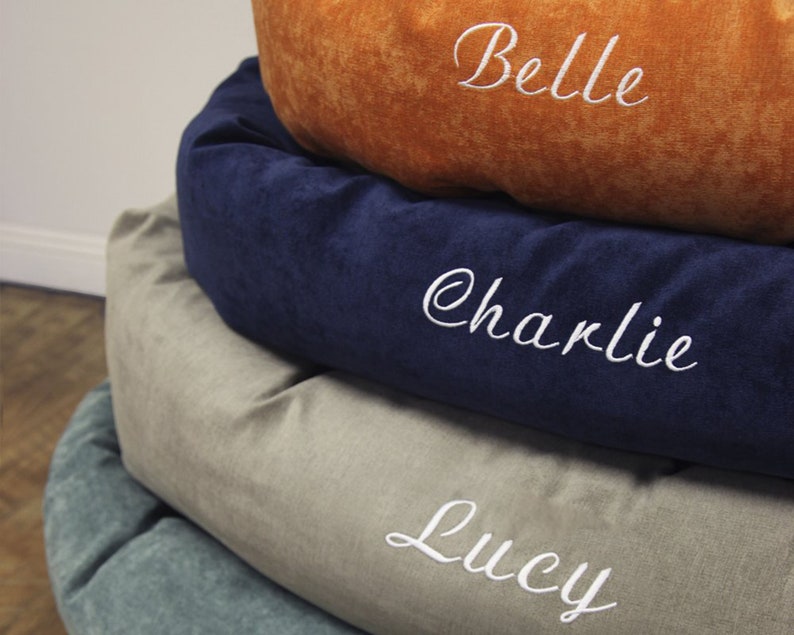 May include: A stack of four round pet beds in different colours and fabrics. The top bed is orange, the second is navy blue, the third is beige, and the bottom is teal. Each bed has a white embroidered name: Belle, Charlie, Lucy.
