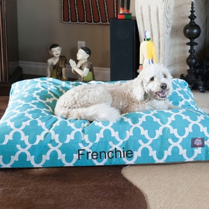 May include: A turquoise and white patterned dog bed with the name "Frenchie" embroidered on the front. A white dog is lying on the bed.