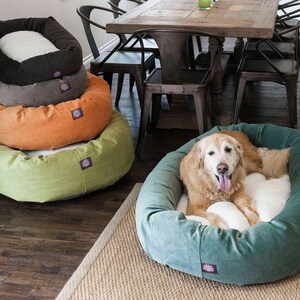 May include: A golden retriever dog relaxing in a teal-coloured dog bed. The bed is round and has a white fluffy inner cushion. The dog is looking directly at the camera with its tongue sticking out. There are four stacked dog beds in the background, in shades of brown, orange, and green.