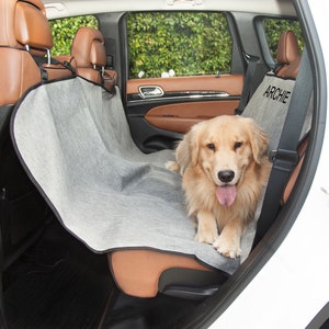 May include: A grey and black dog seat cover with the name "Archie" embroidered on it. A golden retriever dog is sitting on the seat cover in the back seat of a car.