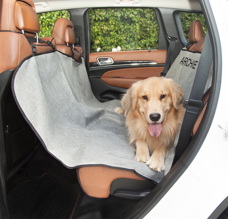 May include: A grey and black dog seat cover with the name "Archie" embroidered on it. A golden retriever dog is sitting on the seat cover in the back seat of a car.