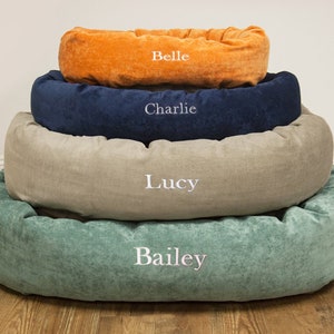 May include: Four dog beds stacked on top of each other. The beds are in different colours and sizes. The top bed is orange and says "Belle". The next bed is blue and says "Charlie". The next bed is beige and says "Lucy". The bottom bed is green and says "Bailey".