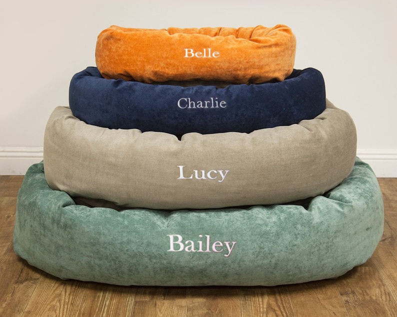 May include: Four dog beds stacked on top of each other. The beds are in different colours and sizes. The top bed is orange and says "Belle". The next bed is blue and says "Charlie". The next bed is beige and says "Lucy". The bottom bed is green and says "Bailey".