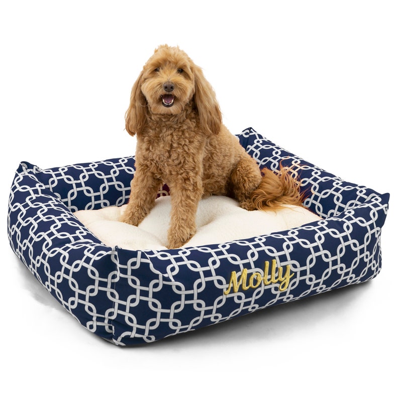 May include: A navy blue dog bed with a white geometric pattern and a gold embroidered name "Molly". A light brown dog is sitting on the bed.
