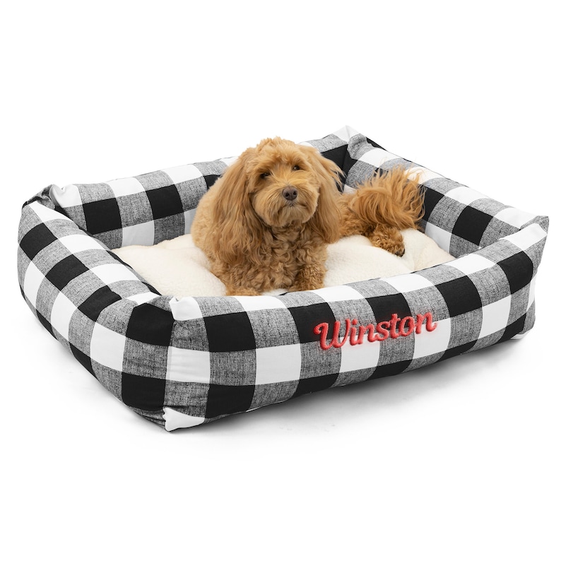Op de afbeelding: Een zwart-witte buffalo plaid hondenbed met een rode geborduurde naam "Winston". Een bruine hond ligt in het bed.