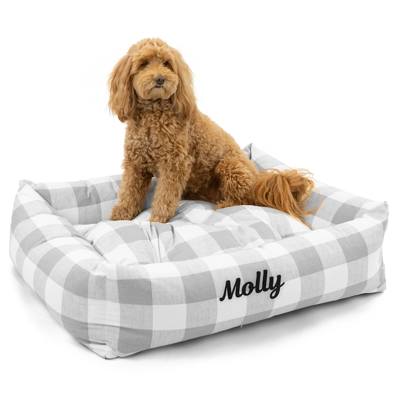 Op de afbeelding: Een grijs en wit geruit hondenbed met een zwarte geborduurde naam "Molly". Een kleine, lichtbruine hond zit op het bed.