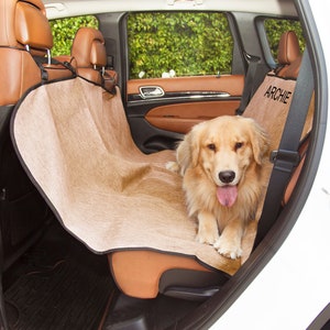 May include: A tan dog seat cover with the name "ARCHIE" embroidered on it. A golden retriever dog is sitting on the seat cover in the backseat of a white car.