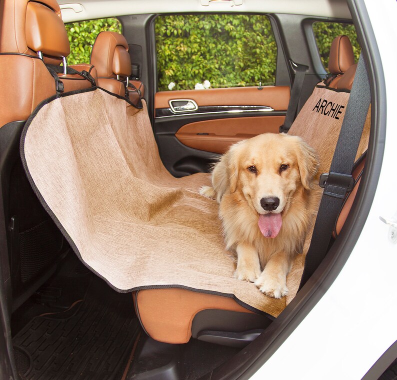 May include: A tan dog seat cover with the name "ARCHIE" embroidered on it. A golden retriever dog is sitting on the seat cover in the backseat of a white car.