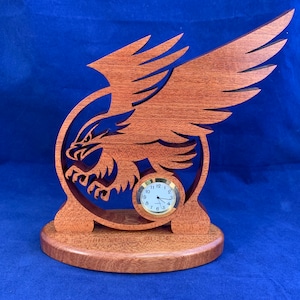 May include: A wooden clock with a carved eagle design. The clock is made of reddish-brown wood and has a round face with black numbers and hands.