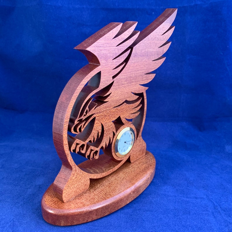 May include: A wooden clock with a carved eagle design. The clock is mounted on a wooden base and has a round face with gold hands and numbers.