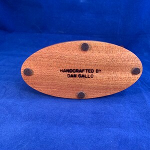 May include: A wooden oval base with four black felt pads on the bottom. The base is engraved with the text "HANDCRAFTED BY DAN GALLO".
