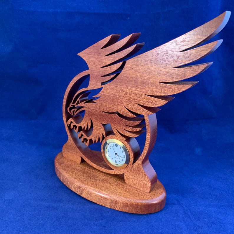May include: A wooden eagle sculpture with a clock face in the center. The eagle is carved in a reddish-brown wood and has its wings spread out.
