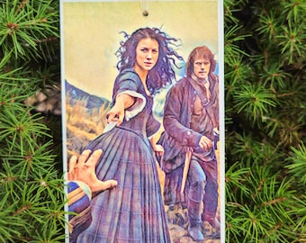 May include: Rectangular ornament featuring a watercolor-style image of two people in period clothing, set against a mountainous backdrop. The woman in a blue plaid dress is reaching out. A ribbon is attached for hanging.