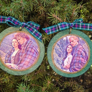 May include: Two round wooden ornaments with green edges, each featuring a printed image of a couple in a plaid shawl. The ornaments are topped with a green, blue, and red plaid ribbon bow for hanging, displaying a romantic scene.