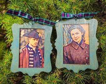 May include: Two wooden ornaments with portraits of people in period clothing. The ornaments have a light blue frame with a decorative edge and are suspended by a plaid ribbon. The portraits are detailed and colorful, set against a background of green pine needles.