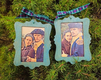 May include: Two teal-colored wooden ornaments with portraits of a couple, each framed by a silhouette. The ornaments are adorned with plaid ribbons and hang against a backdrop of evergreen branches. The portraits feature a man in a blue coat and a woman in a brown coat.