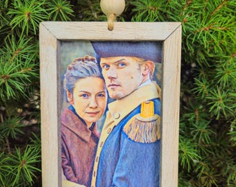 May include: A rectangular wooden framed ornament featuring a portrait of two people. The man is wearing a blue coat with gold epaulettes and a black hat. The woman is wearing a brown coat. The ornament is hanging from a wooden bead and twine.