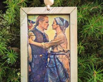 May include: A rectangular wooden framed ornament featuring a historical image of a man and woman in period clothing. The image is set against a backdrop of green pine needles, with a white wooden bead at the top.