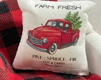 May include: A white pillow with a red truck carrying a Christmas tree. The pillow has the text "FARM FRESH", "PINE-SPRUCE-FIR", "---CUT & CARRY---", and "OPEN DAILY" printed on it. The pillow is sitting on a red and black plaid blanket.