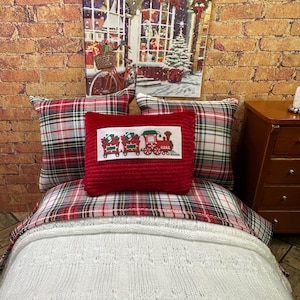 Puede incluir: Una cama de muñecas en miniatura con un estampado de cuadros rojos y blancos. La cama tiene una manta de punto blanca y una almohada roja con un diseño de tren. Una pintura con temática navideña cuelga de la pared de ladrillo detrás de la cama.