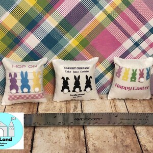 May include: Three miniature pillow cushions with Easter themes. The first pillow is white with pink trim and says "HOP ON" with three cartoon bunnies in purple, blue, and yellow. The second pillow is white with black trim and says "CARROT COMPANY Cake Juice Cookies" with three black bunnies. The third pillow is white with pink trim and says "Happy Easter" with five colorful bunnies.