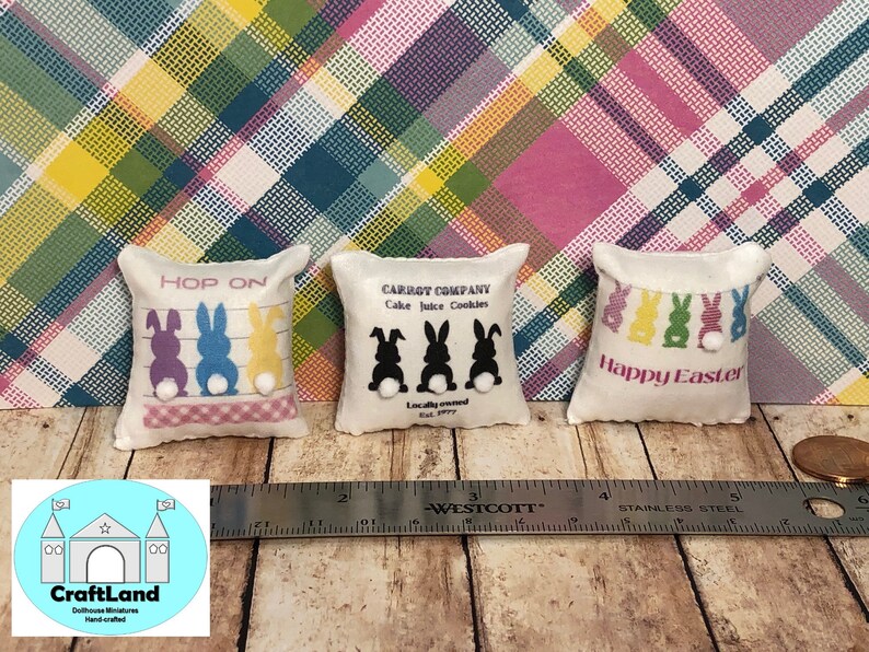 May include: Three miniature pillow cushions with Easter themes. The first pillow is white with pink trim and says "HOP ON" with three cartoon bunnies in purple, blue, and yellow. The second pillow is white with black trim and says "CARROT COMPANY Cake Juice Cookies" with three black bunnies. The third pillow is white with pink trim and says "Happy Easter" with five colorful bunnies.