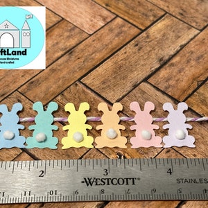 May include: A string of six pastel colored paper bunny cutouts with white pom-poms for tails. The bunnies are strung together with white and purple twine. A ruler is below the bunnies.