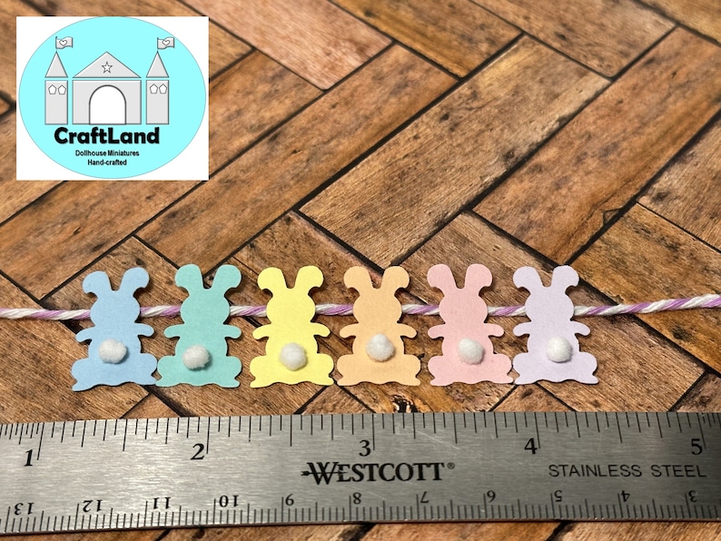 May include: A string of six pastel colored paper bunny cutouts with white pom-poms for tails. The bunnies are strung together with white and purple twine. A ruler is below the bunnies.