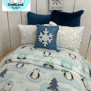 Op de afbeelding: Een miniatuur poppenhuisbed met een wit en blauw dekbed met een pingu&iuml;npatroon, twee witte kussens met blauwe randen en een blauw kussen met een witte sneeuwvlok. Het bed is versierd met een blauw kissen met een witte sneeuwvlok en een bord met de tekst "let it snow".