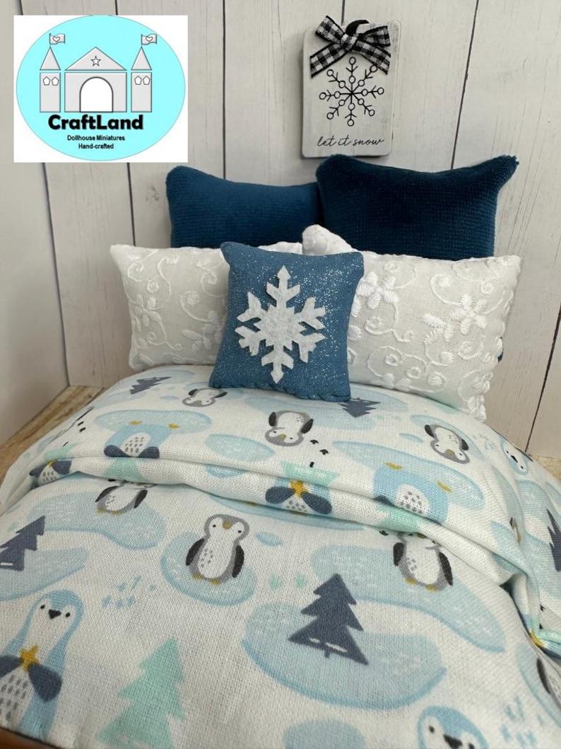 Op de afbeelding: Een miniatuur poppenhuisbed met een wit en blauw dekbed met een pingu&iuml;npatroon, twee witte kussens met blauwe randen en een blauw kussen met een witte sneeuwvlok. Het bed is versierd met een blauw kissen met een witte sneeuwvlok en een bord met de tekst "let it snow".