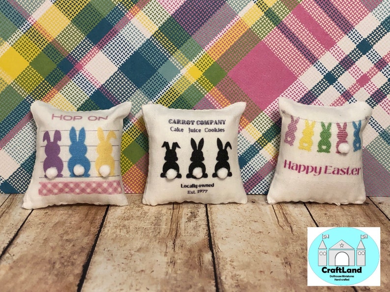 May include: Three small decorative pillows with Easter themes. The first pillow is white with a pink and white checkered border and says "HOP ON" with three bunnies in purple, blue, and yellow. The second pillow is white with black bunnies and says "CARROT COMPANY Cake Juice Cookies Locally owned Est. 1977". The third pillow is white with a pink and white checkered border and says "Happy Easter" with five colorful bunnies.