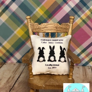 May include: A white pillow with black bunny silhouettes and the text "CARROT COMPANY Cake Juice Cookies Locally owned Est. 1977" on a rocking chair in front of a plaid background.