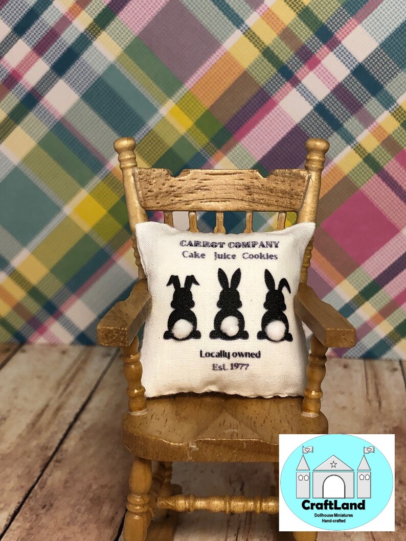 May include: A white pillow with black bunny silhouettes and the text "CARROT COMPANY Cake Juice Cookies Locally owned Est. 1977" on a rocking chair in front of a plaid background.