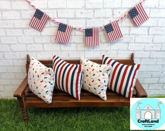 May include: Four decorative pillows with red, white, and blue patterns on a wooden bench. The pillows are arranged in a row, with two striped pillows on the ends and two confetti-style pillows in the middle. The bench is decorated with a string of red, white, and blue American flags.