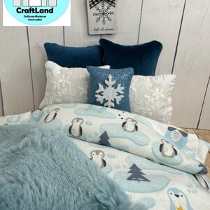 Op de afbeelding: Een poppenhuisbed met een blauwe en witte beddengoedset met een pingu&iuml;npatroon. Het bed heeft een blauwe nep bontdeken en een wit kussen met een sneeuwvlokkenpatroon. Het hoofdeinde heeft een houten bord met de tekst "Let it snow".