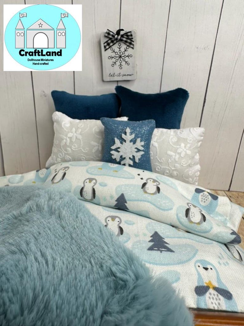 Op de afbeelding: Een poppenhuisbed met een blauwe en witte beddengoedset met een pingu&iuml;npatroon. Het bed heeft een blauwe nep bontdeken en een wit kussen met een sneeuwvlokkenpatroon. Het hoofdeinde heeft een houten bord met de tekst "Let it snow".
