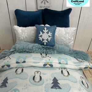 Op de afbeelding: Een miniatuur poppenbed met een blauwe en witte dekbed met pingu&iuml;npatroon, een blauwe nep bont deken en witte en blauwe kussens. Het hoofdeinde heeft een houten bord met een sneeuwvlok en de tekst "let it snow".
