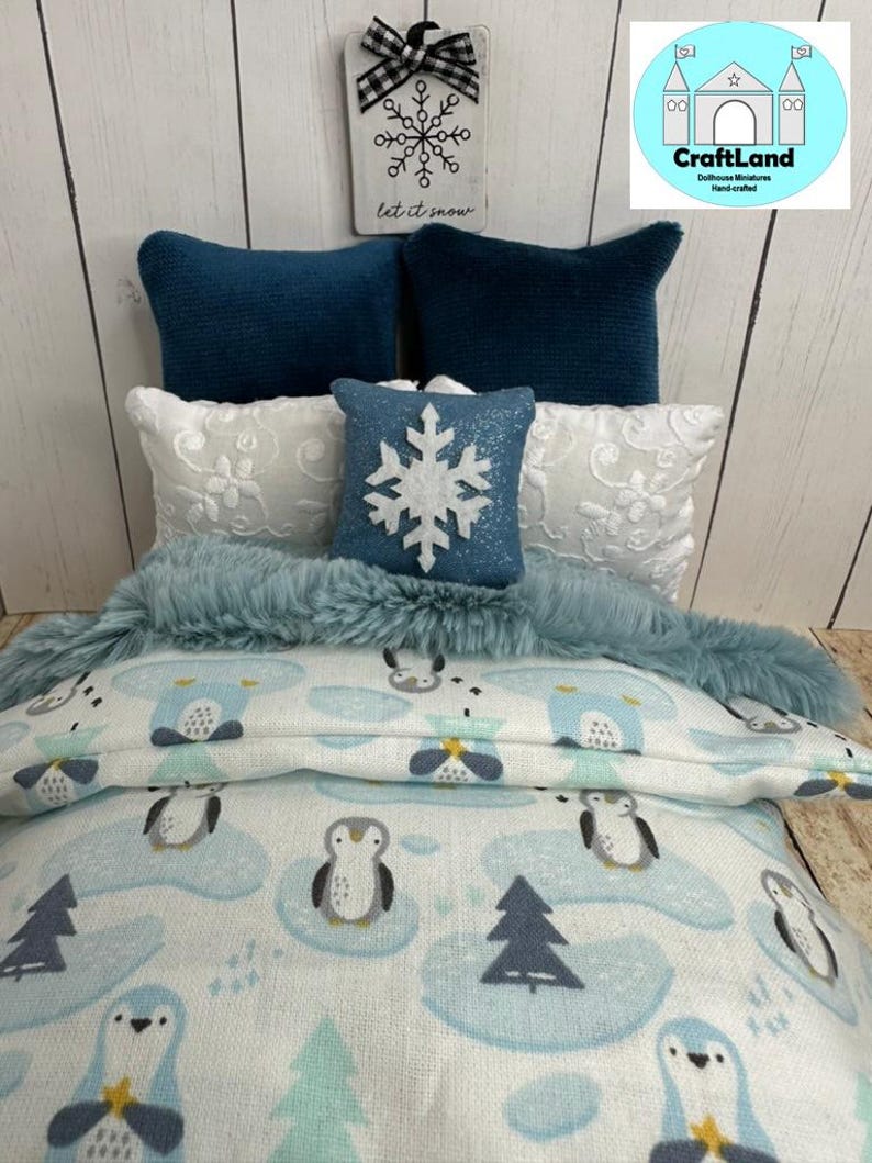 Op de afbeelding: Een miniatuur poppenbed met een blauwe en witte dekbed met pingu&iuml;npatroon, een blauwe nep bont deken en witte en blauwe kussens. Het hoofdeinde heeft een houten bord met een sneeuwvlok en de tekst "let it snow".