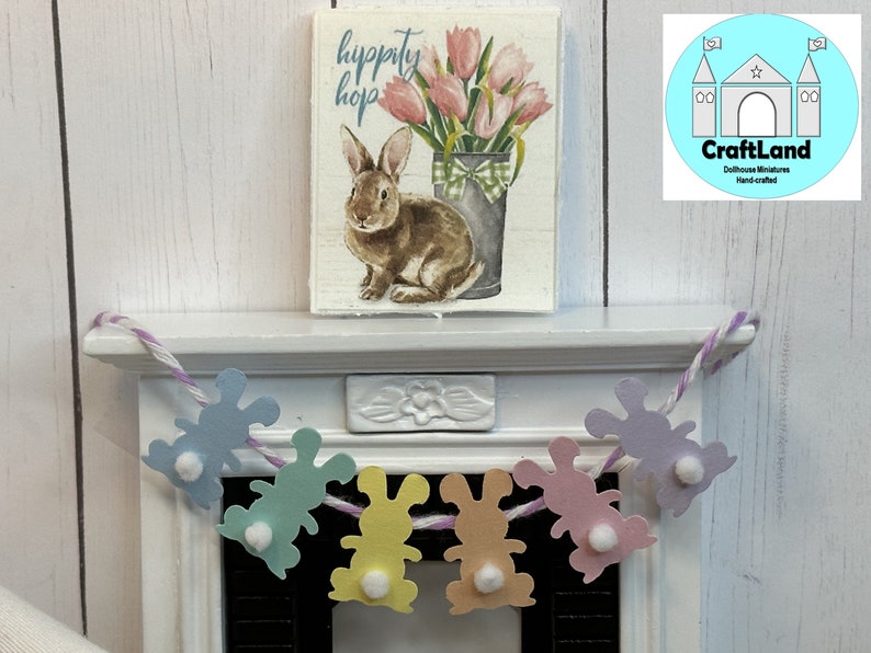 May include: A pastel colored felt bunny garland with white pom-poms hangs on a white mantel. The garland says "hippity hop" and is perfect for Easter decor.