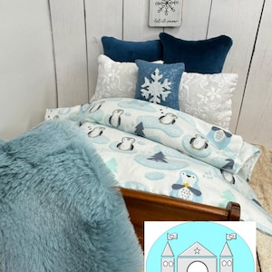 Op de afbeelding: Een poppenbed met een blauwe en witte beddengoedset met een pingu&iuml;npatroon. Het bed heeft een blauwe nep bontdeken en een wit kussen met een sneeuwvlokkenpatroon. Het hoofdeinde is van hout en heeft een wit bord erboven met de tekst "Let it snow".