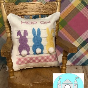 May include: A white pillow with three bunny silhouettes in purple, blue, and yellow with white pom-poms for tails. The pillow has the text "Hop On" printed on it. The pillow is sitting on a wooden rocking chair.