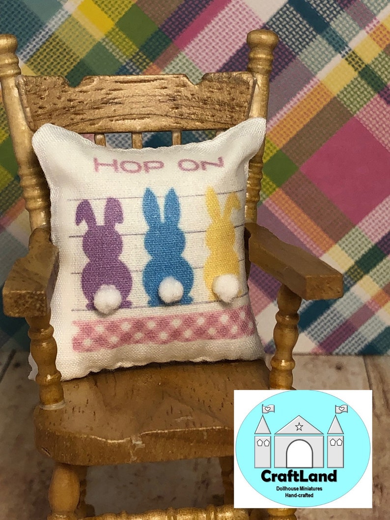 May include: A white pillow with three bunny silhouettes in purple, blue, and yellow with white pom-poms for tails. The pillow has the text "Hop On" printed on it. The pillow is sitting on a wooden rocking chair.